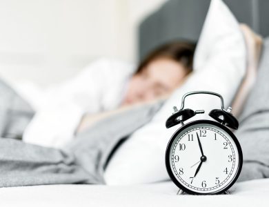 A retro alarm clock sits by a sleeping woman in a cozy bedroom setting.