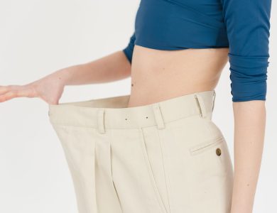Unrecognizable slim female with bare belly wearing white oversized pants after weight loss on white background in modern light studio