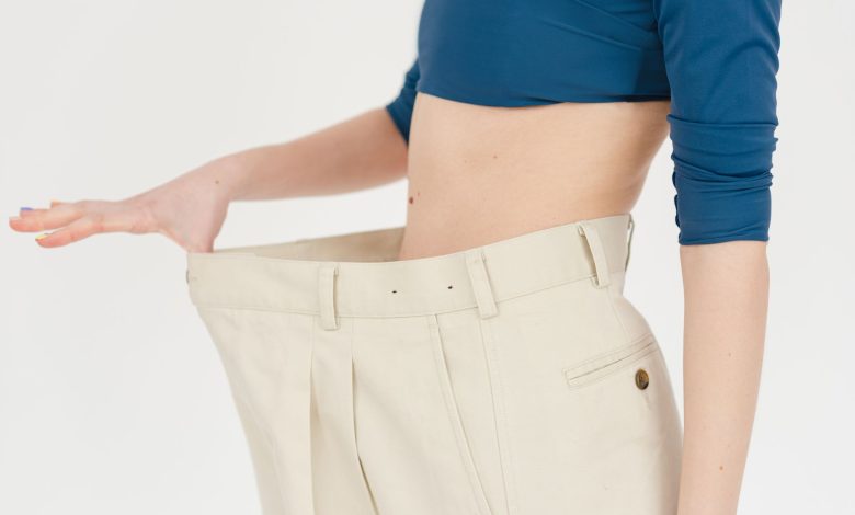 Unrecognizable slim female with bare belly wearing white oversized pants after weight loss on white background in modern light studio
