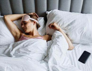 Woman resting in bed wearing an eye mask with a smartphone nearby, embracing relaxation.