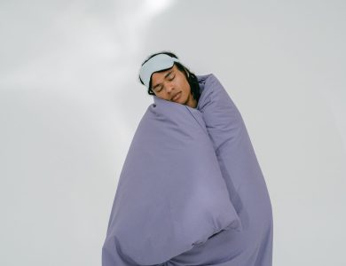 A person peacefully sleeping with an eye mask, wrapped in a purple blanket on a white background.