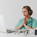 Female nurse using telemedicine for a virtual consultation on a laptop, wearing headphones and stethoscope.
