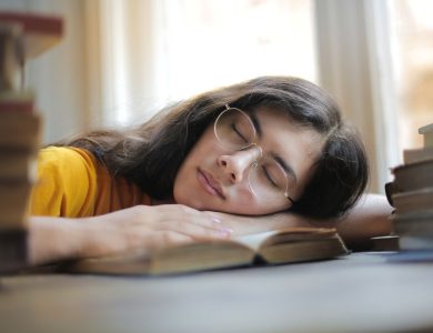 A young woman with glasses naps on a book indoors during the day, capturing a moment of relaxation and fatigue.