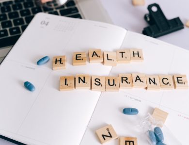 Scrabble tiles spelling 'Health Insurance' on planner with pills and laptop, symbolizing healthcare planning.