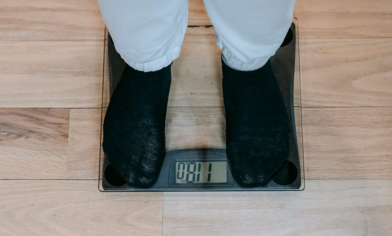 From above of crop unrecognizable plus size person in casual clothes standing on weighing scale on wooden floor