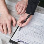 Close-up image of two people signing an insurance policy document on a wooden desk.