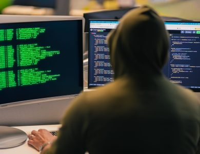 A person in a hoodie coding on dual monitors, depicting cybersecurity and hacking themes.