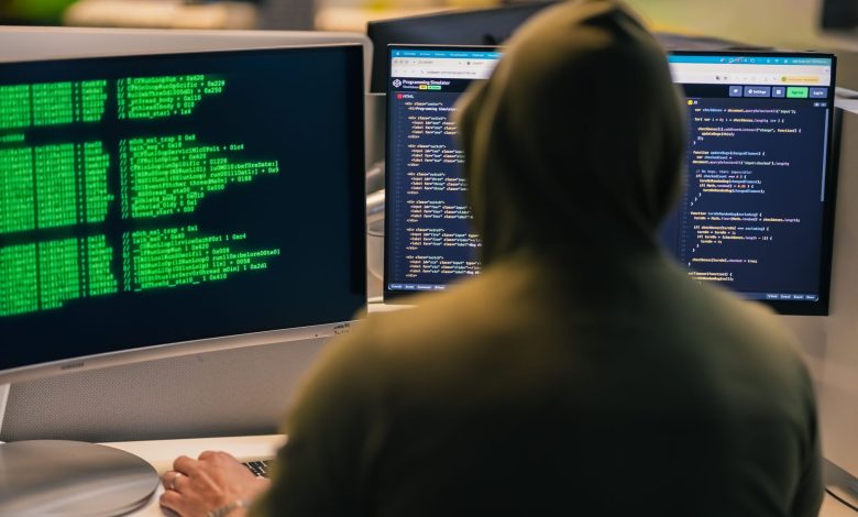 A person in a hoodie coding on dual monitors, depicting cybersecurity and hacking themes.