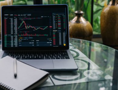 Laptop displaying cryptocurrency stocks and graphs on a glass table with a notepad.