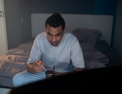 Adult male enjoying a dessert watching TV in a cozy bedroom setting.