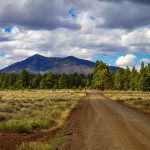 A picturesque dirt road in Flagstaff, Arizona with mountains and lush greenery under a cloudy sky.