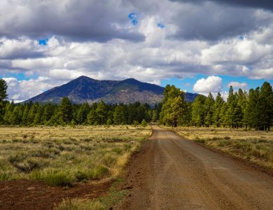 A picturesque dirt road in Flagstaff, Arizona with mountains and lush greenery under a cloudy sky.