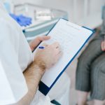 Medical professional writing patient notes on clipboard during consultation.