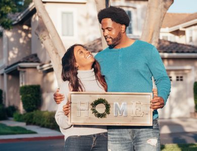 Joyful couple celebrating home purchase, holding 'Home' sign outdoors.