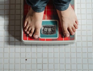 From above crop anonymous barefoot child in jeans standing on weigh scales on tiled floor of bathroom